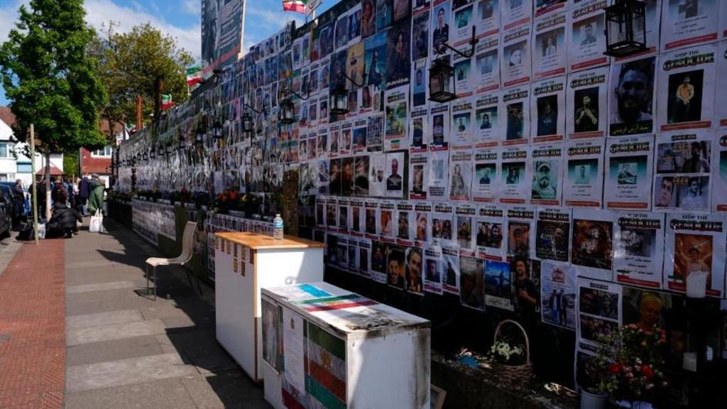 A wall of people's faces and information is topped by Iranian flags. Flowers and furniture also line the wall.