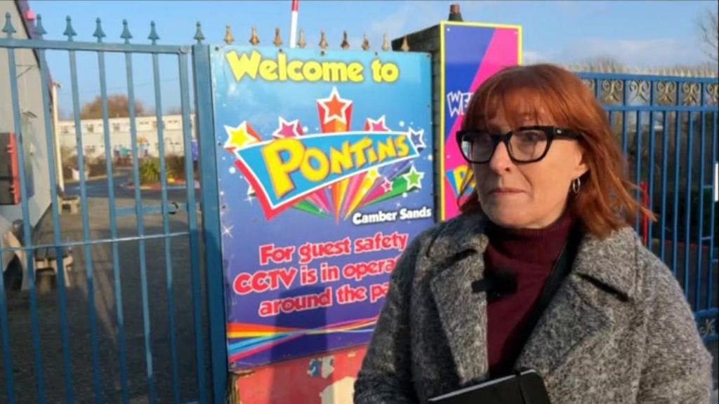 A woman with shoulder length red hair and thick black glasses stands outside the blue iron gates of the Pontins holiday park in Camber
