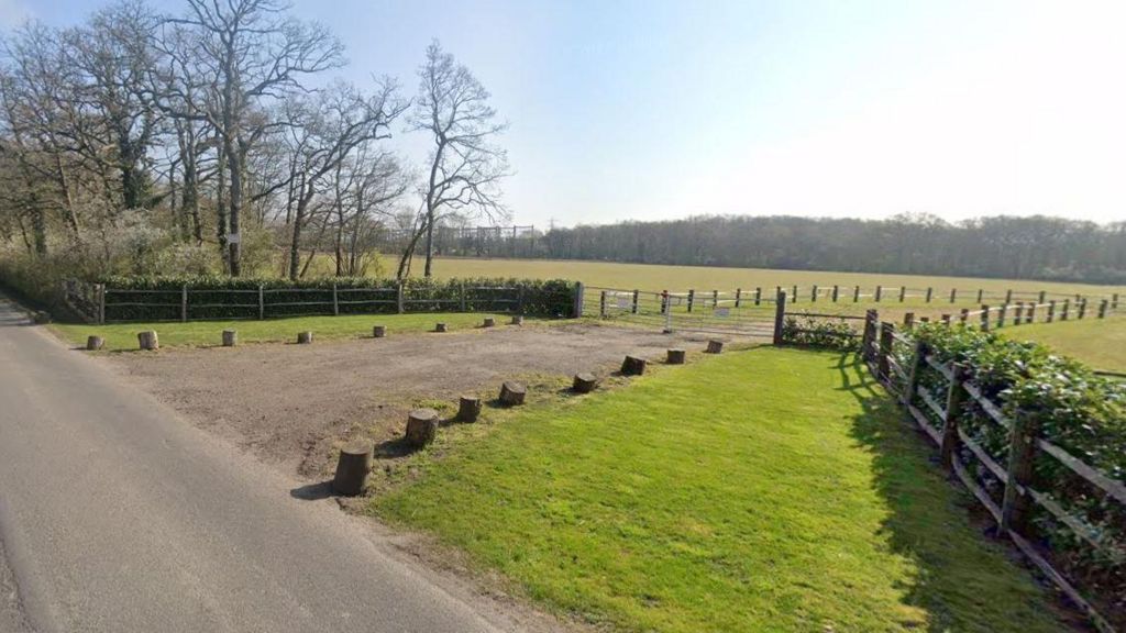 A Google maps image of a dirt driveway up to a large green field enclosed by wooden fencing and mature trees 