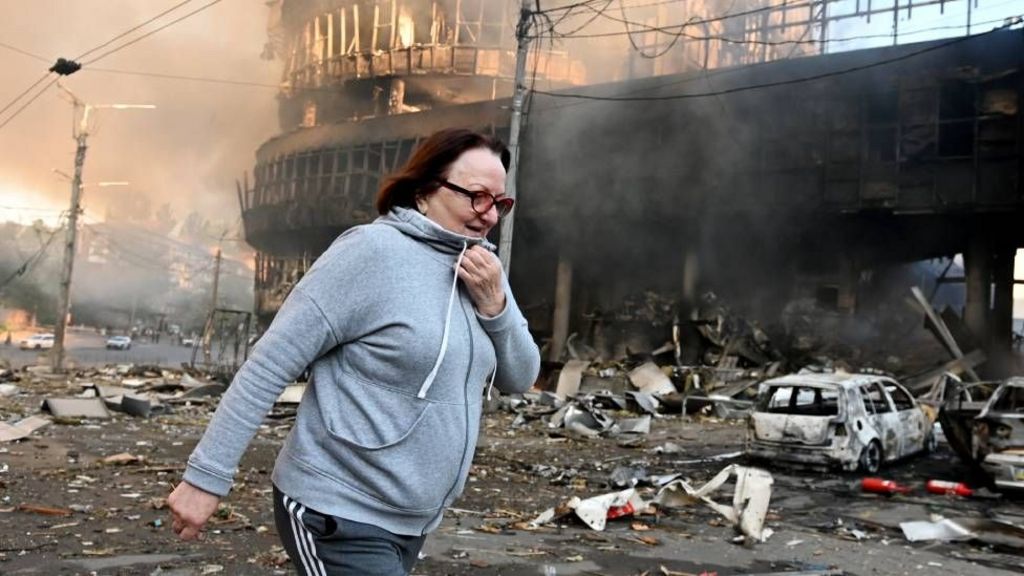 A woman in a grey hoodie and track pants walks past a destroyed building in Kyiv on May 24. There is debris on the floor and smoke in the air. The charred remains of a car can be seen in the background