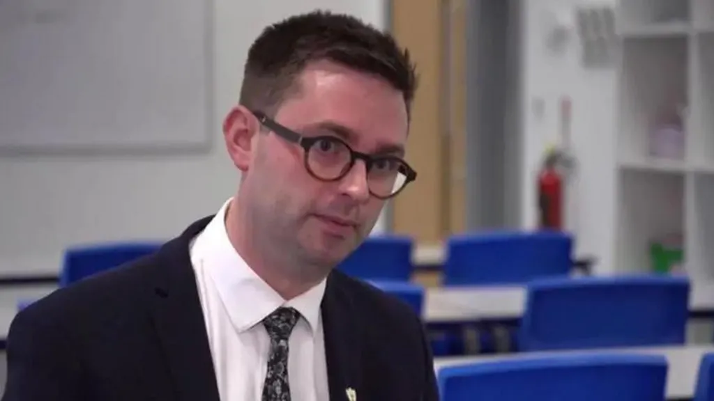 A man with short black hair is wearing black rimmed glasses, a black suit and a white shirt with dark tie. He is standing in a school classroom