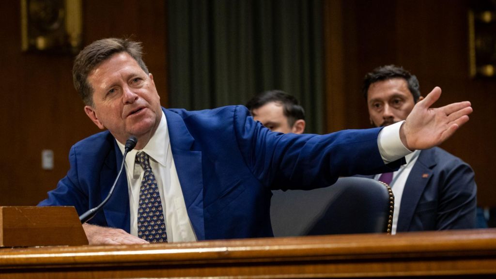 Jay Clayton sitting at a bench inside the Dirksen Senate Office Building in Washington, leaning into a microphone and stretching his arm out