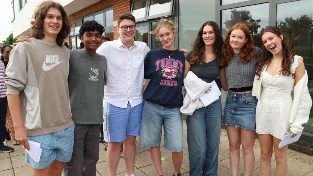 Students collecting their A Level results at Pate's Grammar School. Seven students are standing in a line with their arms linked and smiling at the camera, wearing casual clothes