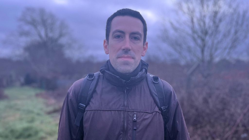 A man with dark hair and a brown jacket stands with a black backpack on. In the background is a green area with trees.