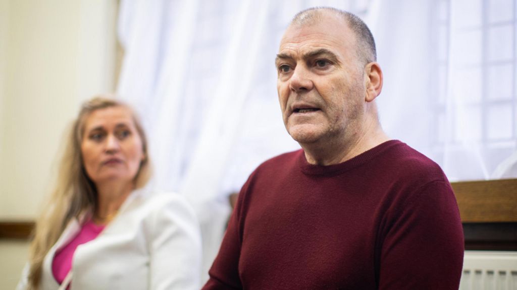 Mark Hehir speaking while seated at a table, with Katalin Kaszas sitting behind him and looking on.