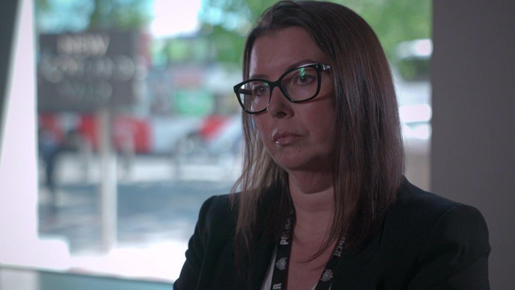 A middle-aged woman is sitting in a grey room, in front of a window. Outside, the New Scotland Yard sign can be seen. She has long dark hair and wears glasses and a black suit jacket. 