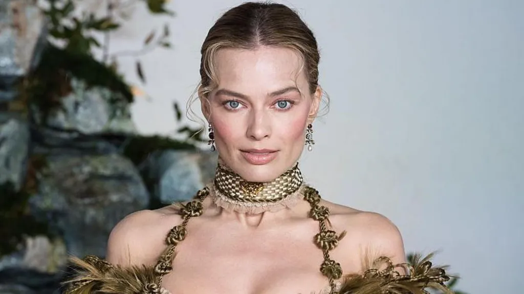 Margot Robbie has her blonde hair swept back with two curly tendrils at the front. She has dangly earrings and a gold choker-style thick necklace