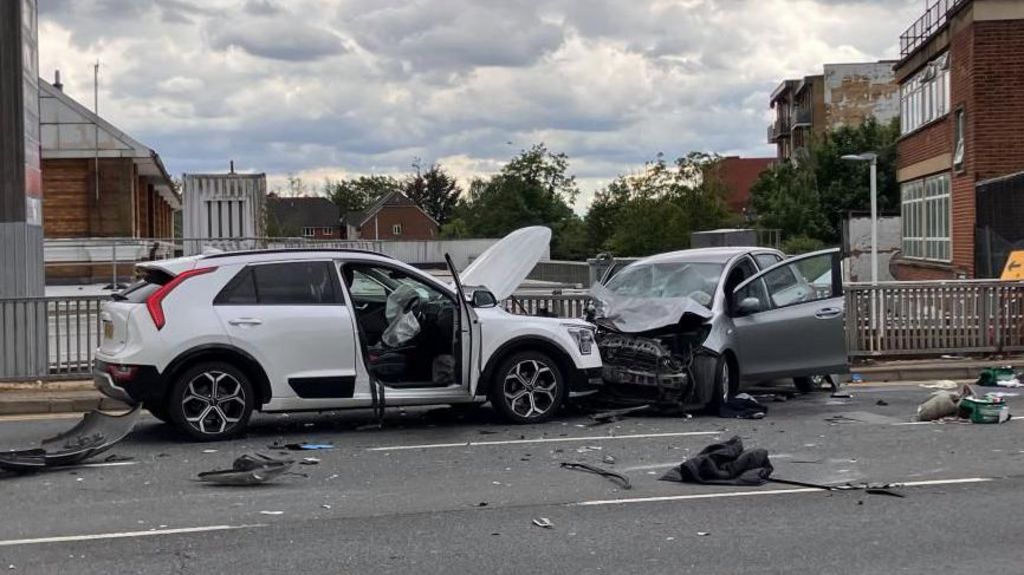 A white car and a grey car are badly damaged from the impact with debris scattered across the road.