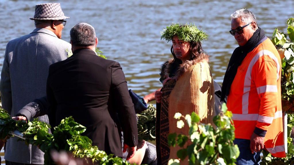 Maori Queen Kuini Nga wai hono i te po stands next to a river wearing a headress and a cloak, with three men standing alongside her.