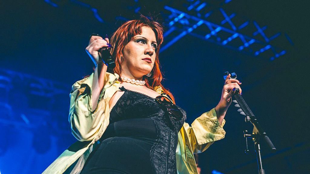 CMAT is performing on stage and holds a microphone in one hand and a microphone hand in the other. She wears a black silk yellow shirt over a black corset top. She has ginger hair.