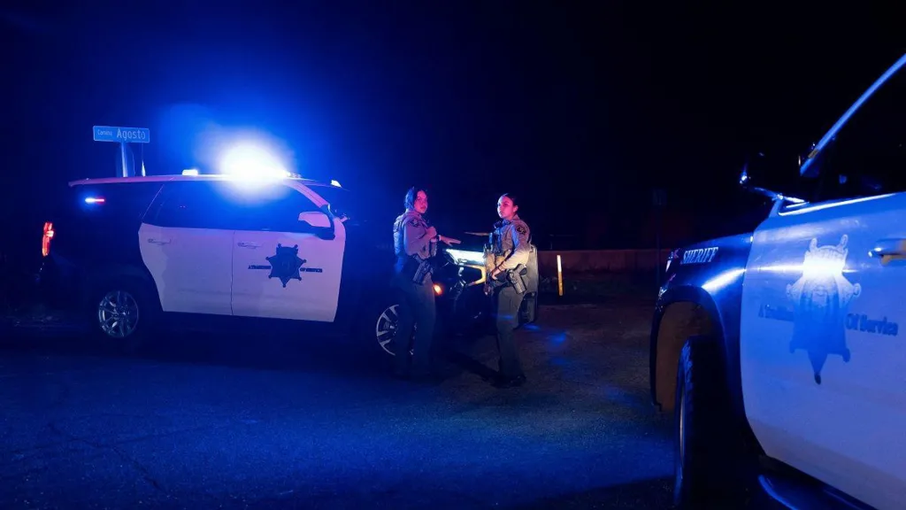 Two female police officers in uniform stand in front of a police vehicle, with its headlight turned on in the night.