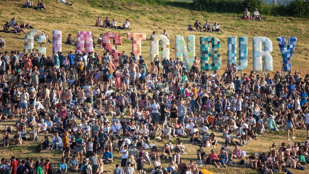 Hundreds of people gathered, standing and sitting, on the grassy hill beneath the colourful Glastonbury sign. 