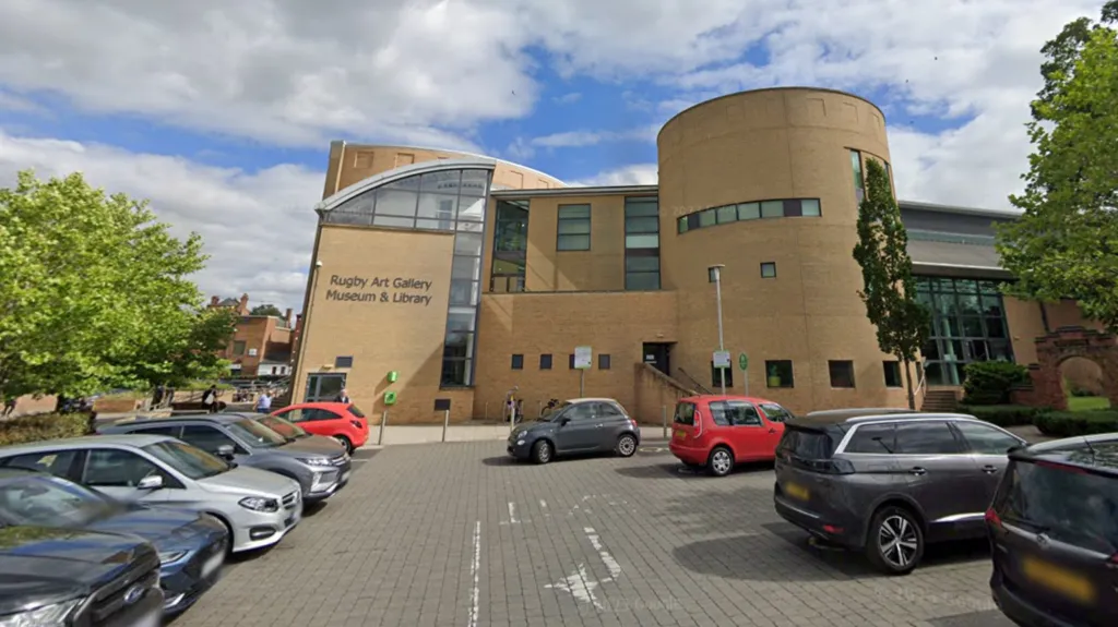 A modern, light-coloured brick building with lettering on the side which indicates it houses an art gallery, museum and library. There is a car park in the foreground of the image with car parks in rows on either side. There are a number of trees nearby the building.