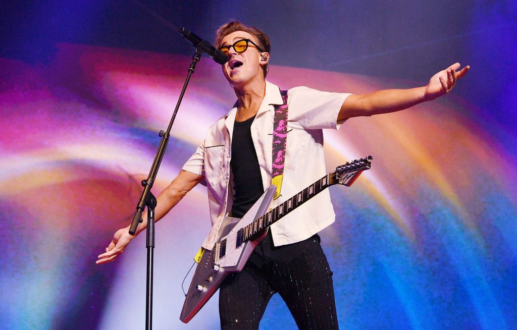 Tom Fletcher holds his arms wide open as he sings into a microphone with a 'flying V' style guitar slung around his neck and shoulders, wearing a white shirt and black trousers against a colouful background during the Busted vs McFly tour at Wembley in September.