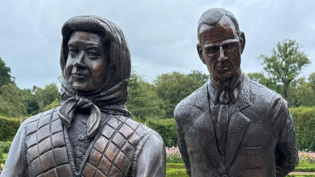 The bronze sculpture of the royal couple and their two corgis in the Parterre Garden at Antrim Castle Gardens, surrounded by flowers and under a grey looking sky with trees in the background.   The late queen (sculpture) is wearing a cardigan, scarf and carrying a handbag. She is holding her hands together.   The late king (sculpture) is wearing a suit.   The corgi dogs are sitting down, one is looking up at the queen and one is looking ahead.