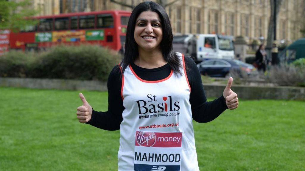 A woman with long black hair smiling, in a white vest and a London Marathon number on it