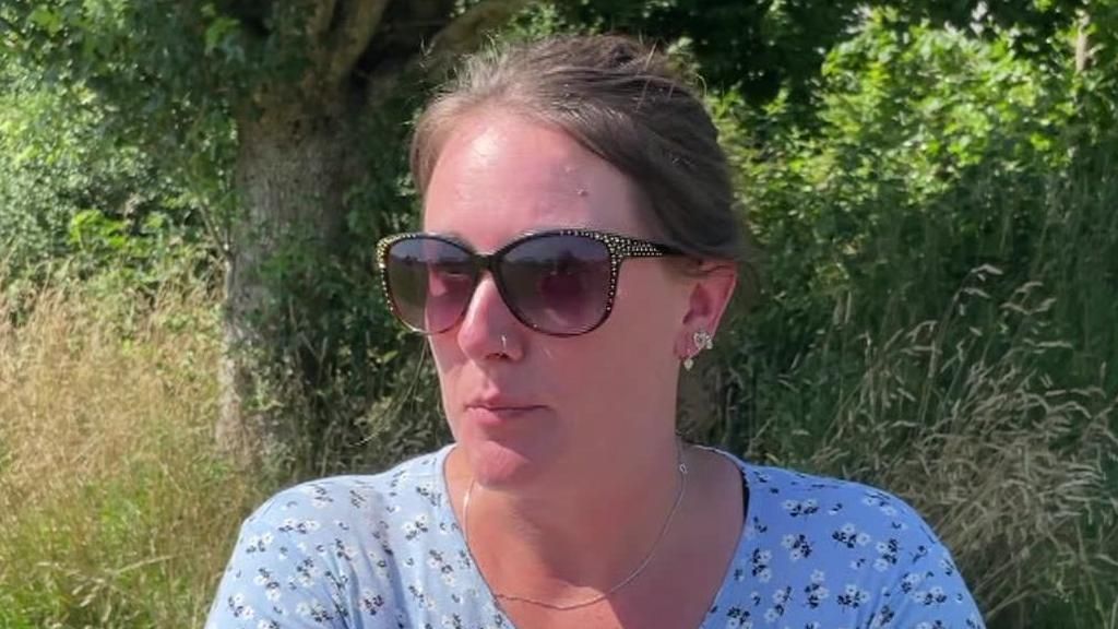 Head shot of Sherford resident Lindsay Britain were sunglasses and a blue dress with white flowers on it stood in front of a tree and grass verge.