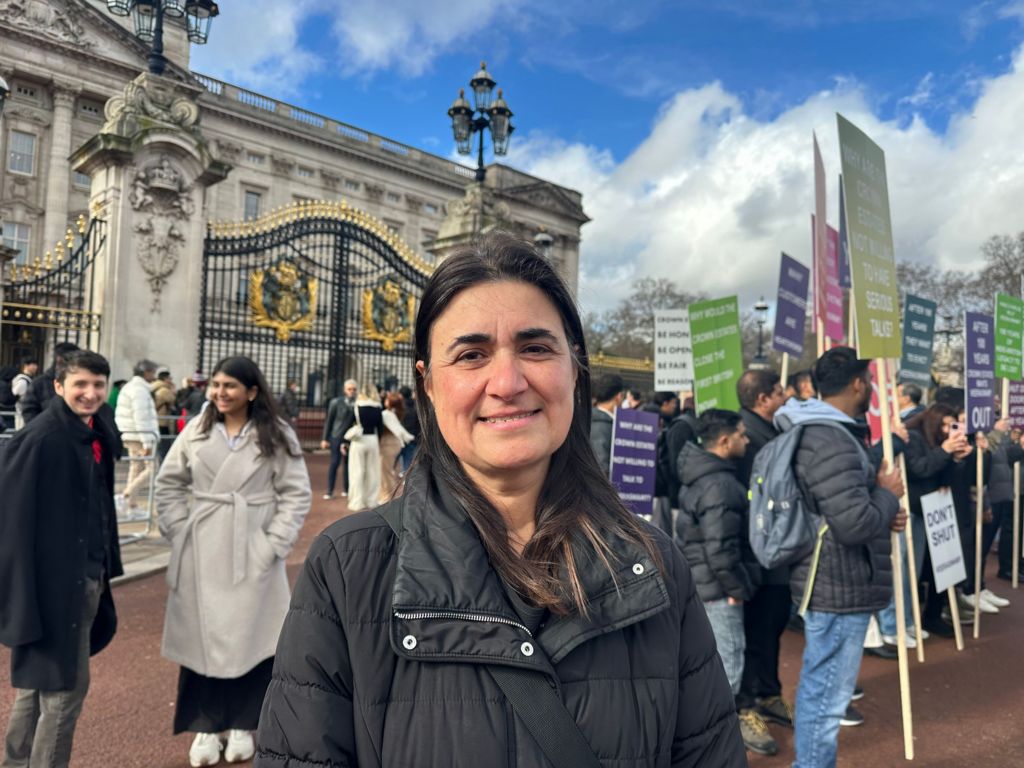 A woman in a black coat standing to the side of the protest outside Buckingham Palace.