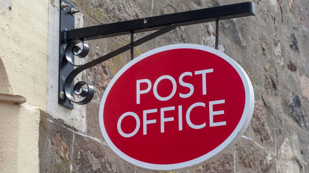 A red and white Post Office sign attached to a historic stone wall being held by a black metal beam
