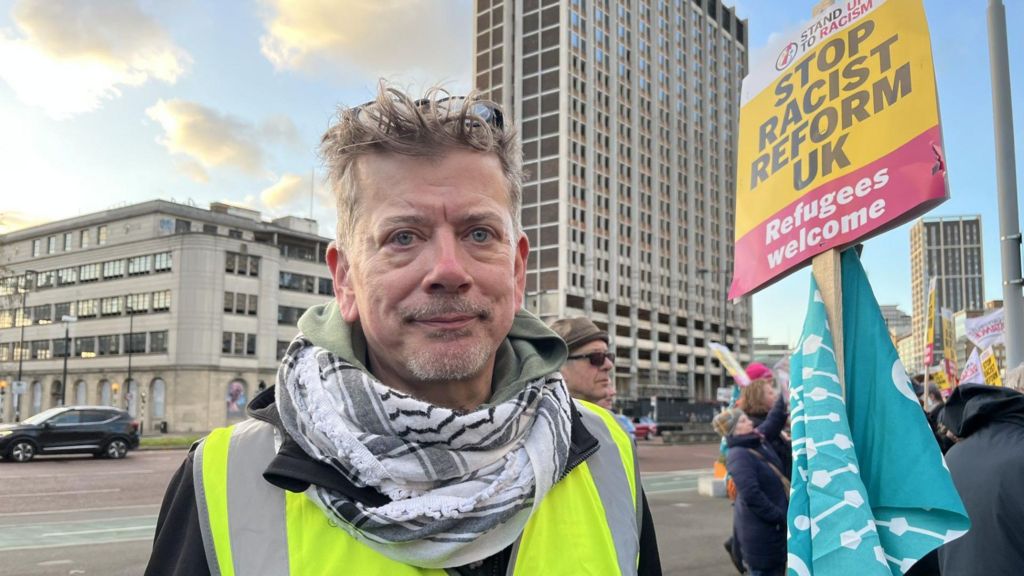 Michael Holland facing the camera wearing a high visibility vest and black and white scarf. He has short blond spikey hair and is surrounded by protesters and is holding up a sign that says 'Stop Racist Reform UK'.