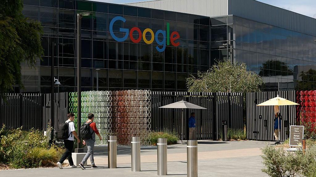 Outside of a large dark-coloured shiny building with the Google logo in blue, red, yellow and green letters high up on the wall. In front of it two young unidentifiable men with large backpacks are walking with their backs to us