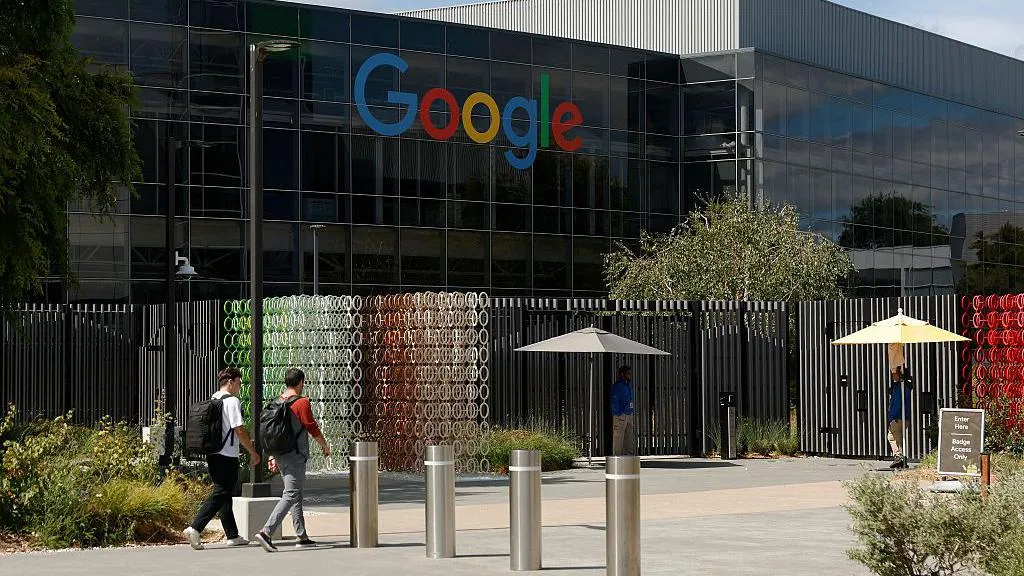 Outside of a large dark-coloured shiny building with the Google logo in blue, red, yellow and green letters high up on the wall. In front of it two young unidentifiable men with large backpacks are walking with their backs to us