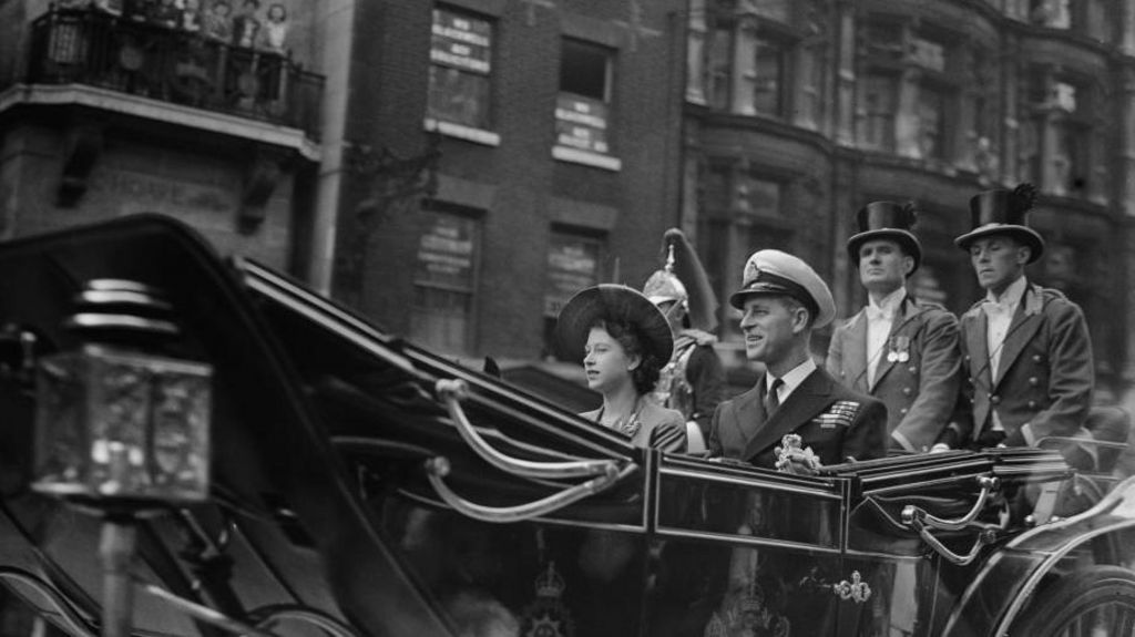Princess Elizabeth (later Queen Elizabeth II, 1926 - 2022) sitting in an open carriage accompanied by her husband Prince Philip who is wearing a uniform with a peaked cap - there are two coachmen sitting behind. 