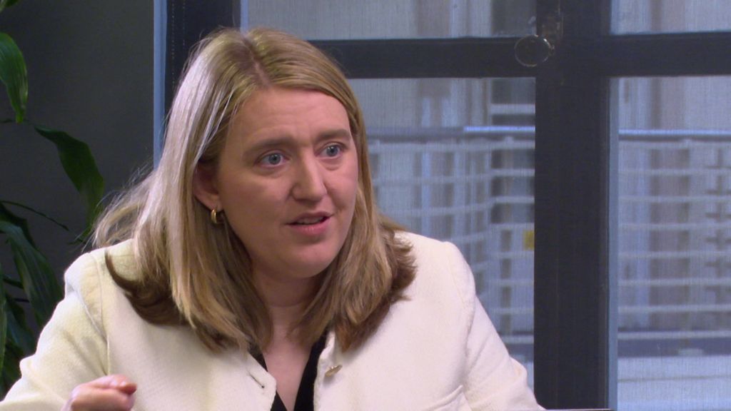 Woman with light blondish hair, wearing a white jacket and black top underneath 