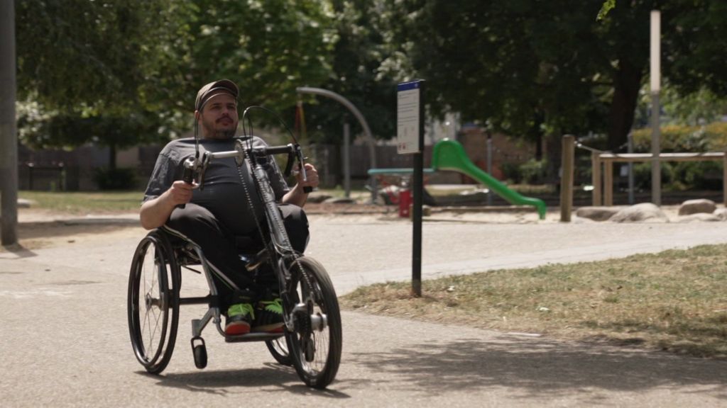 A man is in a wheelchair in a park. He is using a hand cycle to propel himself.