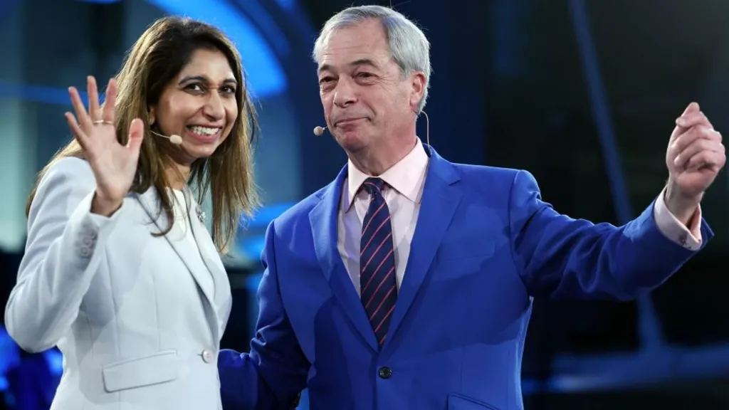 Suella Braverman waves with her right hand as she grins and moves towards Nigel Farage who smirks as he holds aloft his left arm. Both are wearing blue suits and have small headset microphones.