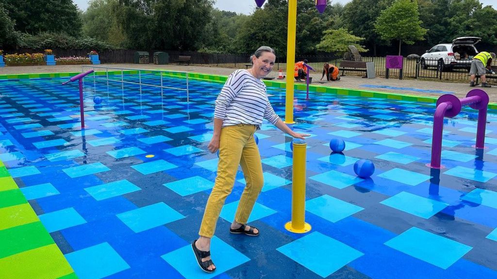 A woman wearing a stripy black and white top, yellow trousers and black sandals rests her hand above a water feature in a splash park