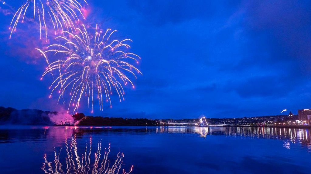 fireworks go off above the river Foyle , the craigavon bridge is in the background. It is dark and thebridge is lit up  