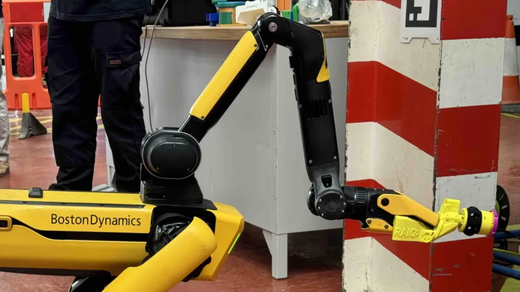 A four-legged, yellow and black robot with Boston Dynamics written on its side. It has a large, extendable arm at the front which holds a yellow swabbing tool.