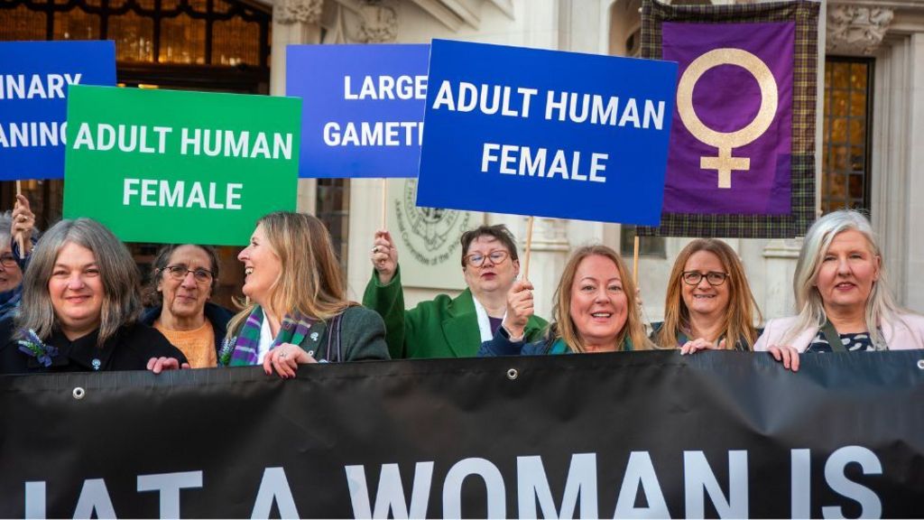 Women line up behind a black banner being held in front of them. Some are also holding signs above their heads, including two reading "adult human female". 