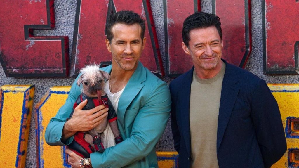 Ryan Reynolds holds a dog in a Deadpool outfit next to Hugh Jackman on the red carpet