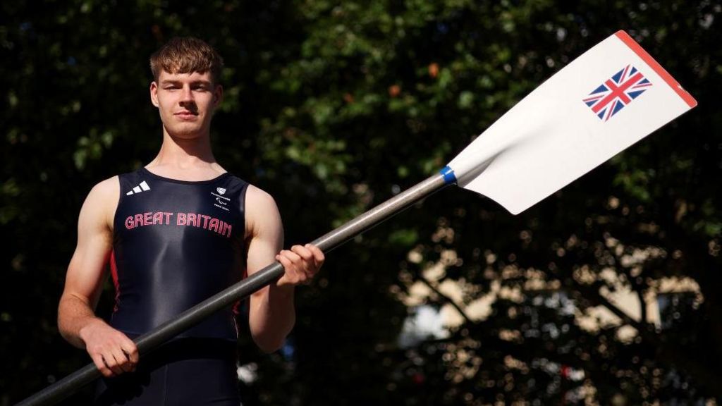 A man in a navy sports Team GB one-piece holds up a white oar with the Union Jack flag on it. He is looking directly at the camera and photographed outside. 