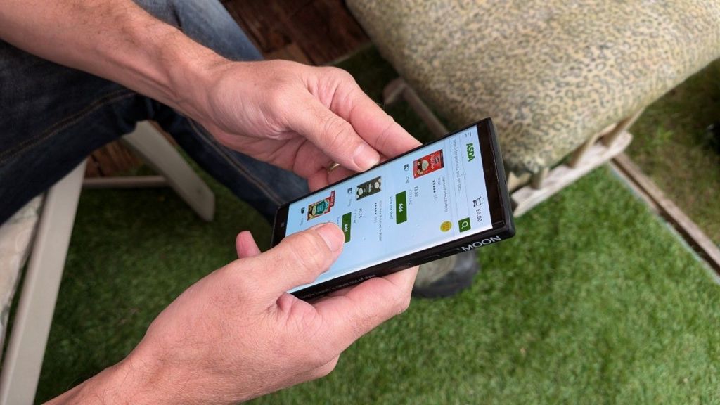 Mr Windell holding a black phone with the screen displaying items from Asda's online shopping range