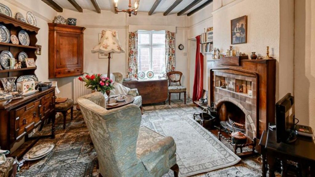 A living room with a brick fireplace and two armchairs, as well as a sideboard with shelves to display numerous items of crockery