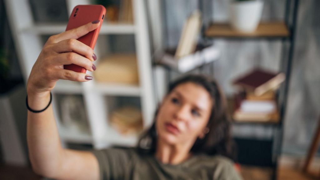 A woman in a dark green T-shirt is shown sitting in a chair, with her arm raised above her head as she holds her phone to take a selfie.