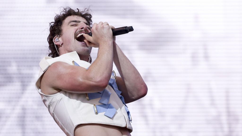 Benson Boone, wearing a white waistcoat with powder-blue military style detail, sings passionately into a microphone with his head tilted back and eyes closed.