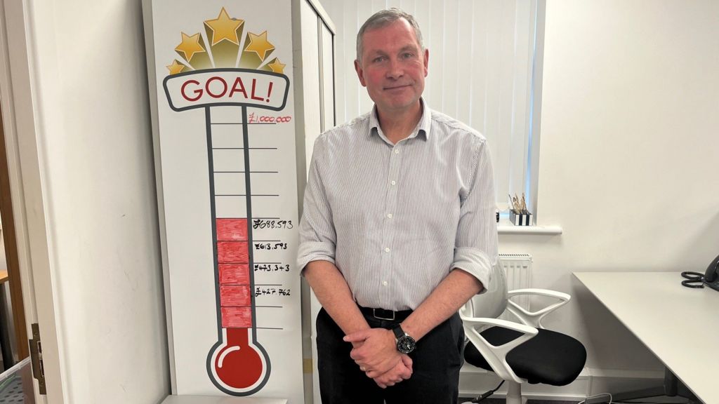 Dave Mead, head of the Victim Care and Support Service standing next to a wall chart showing how much money they've managed to get refunded to fraud victims through their advocacy work