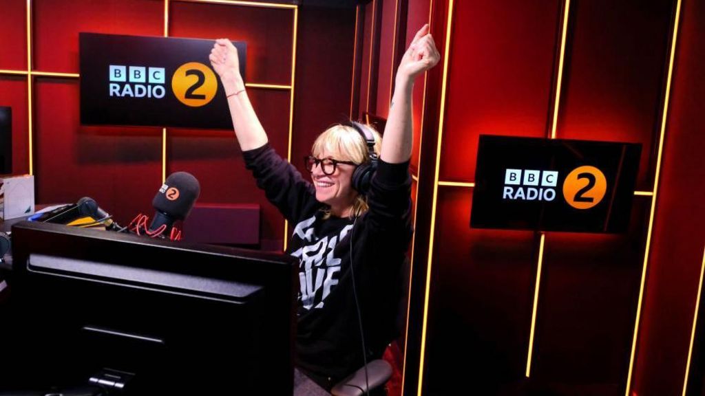 Zoe Ball seen in the BBC Radio 2 studio smiling with her arms raised.