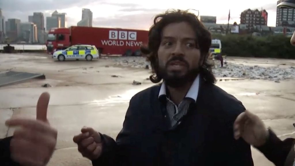 A man with long dark hair, a beard and wearing a jacket stands with a fake police car and fake BBC World News lorry in the background