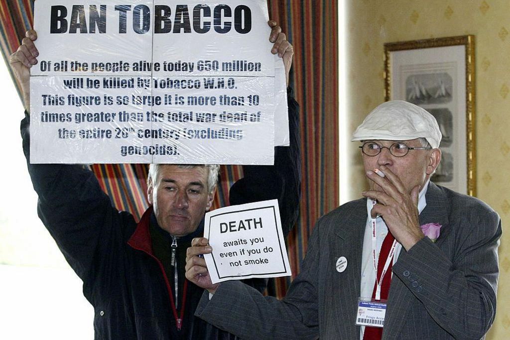 David Hockney and an anti-smoking protester in 2005. The protester is holding up a sign calling for tobacco to be banned. Hockney is holding up a small counter sign saying that people die even if they don't smoke. He is smoking.