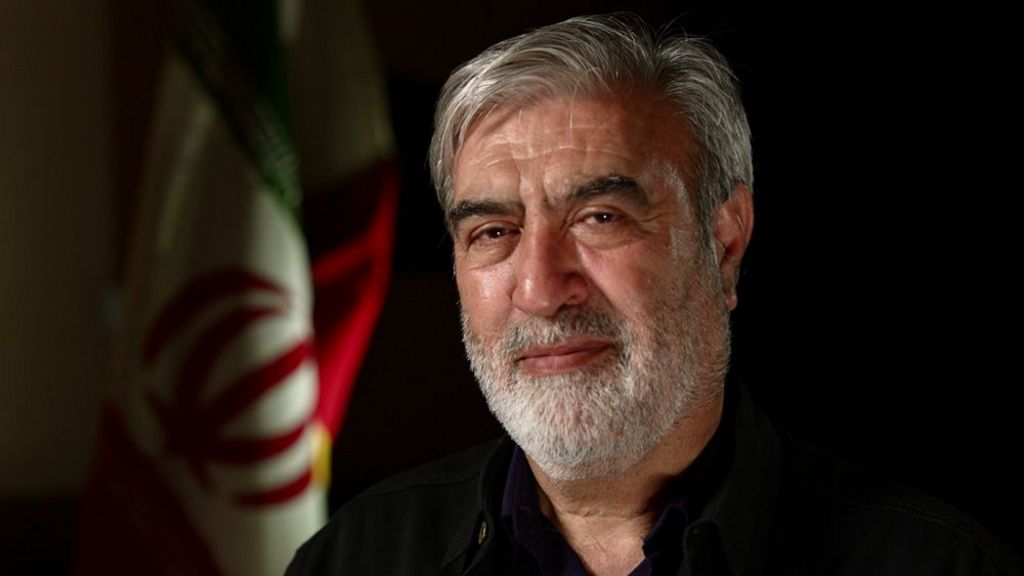 A man with a grey beard and hair looks into the camera. He is wearing a black shirt and behind him is the Iranian flag.