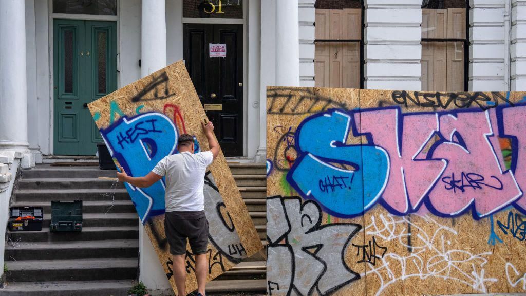 The large decorative chipboard panels being put up in front of a row of terraced houses. One house is already covered and just behind one of the chipboards the man is carrying, you can see the steps leading up to a house and the two front doors, one black, one green.