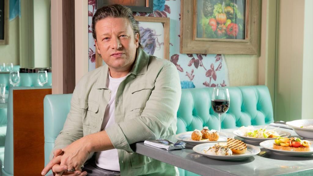 Jamie Oliver is sitting on a banquette at a table covered with four plates of food and a glass of red wine. He is wearing a casual shirt over a white t-shirt and looks serious but not unhappy.