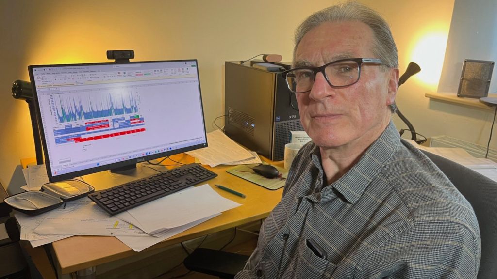 A man with short gray hair and glasses, wearing a gray patterned shirt, sitting at a desk. He looks directly at the camera with a neutral expression. In the background, a computer monitor displays a graph with red and blue lines, along with a keyboard, mouse, papers, and a computer tower.