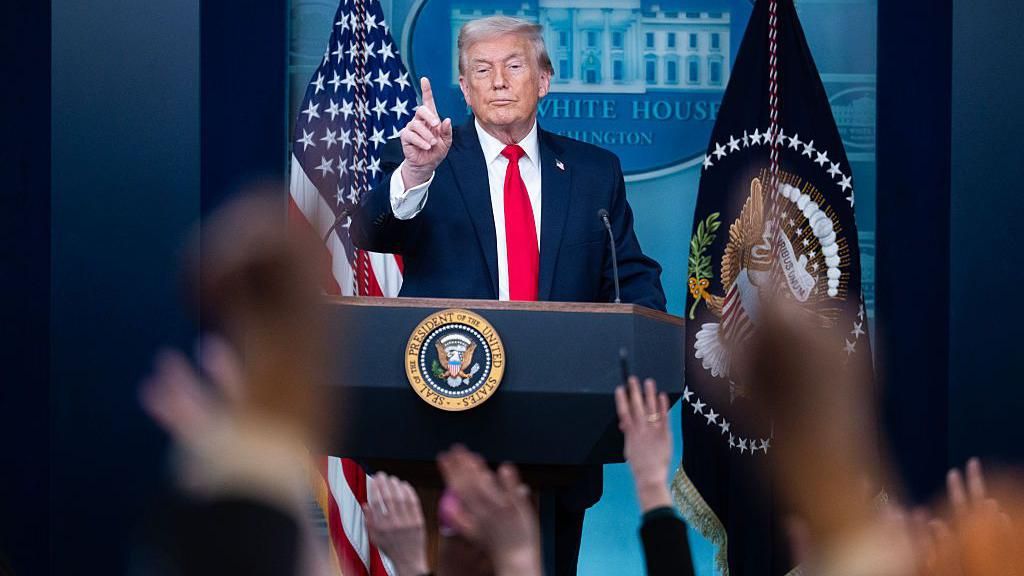Donald Trump at a White House briefing with journalists hands up to ask a question in the foreground