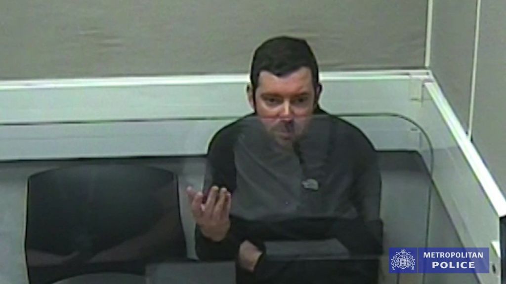 Scott O'Connor in a police interview. He is wearing a black top and sitting behind a transparent screen in a room with grey walls. 